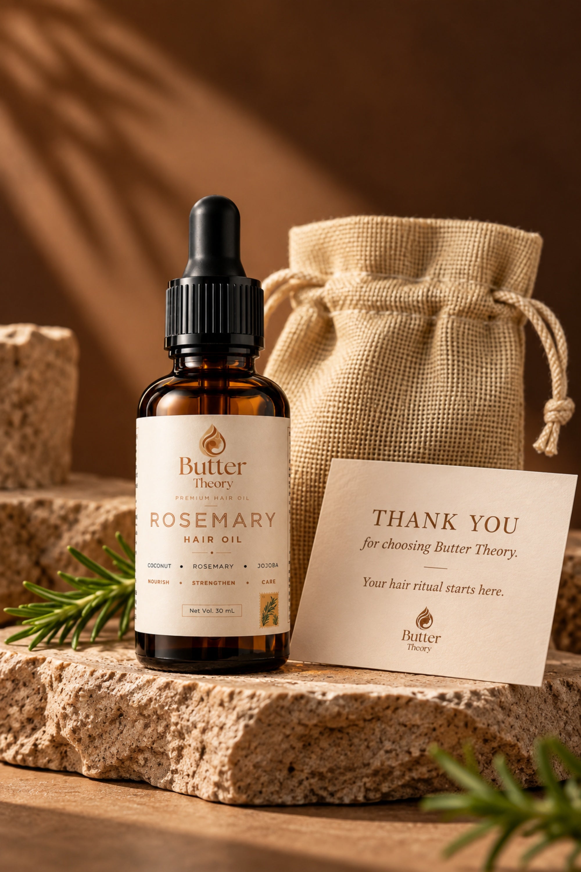 Butter Theory Rosemary Hair Oil in an amber glass dropper bottle, styled on a rustic stone slab with a burlap drawstring pouch and a thank you card in the background.